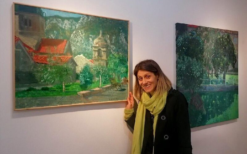 Изображение женщины на выставке картин в Черногории, показывающей на пейзажи с красной крышей и зеленью.