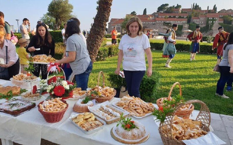 Женщины на фестивале в Черногории выставляют традиционные блюда на столе на фоне исторической архитектуры.