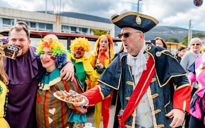 Карнавальное мероприятие в Черногории: участники в красочных костюмах и клоуны с едой на улице.