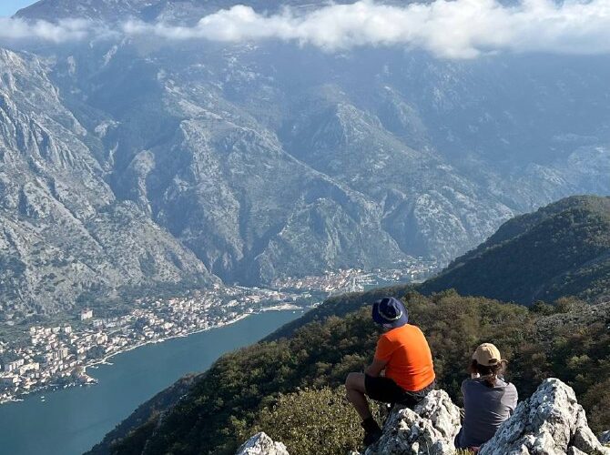 Два туриста любуются пейзажем Которского залива в Черногории, сидя на скалах в солнечный день.